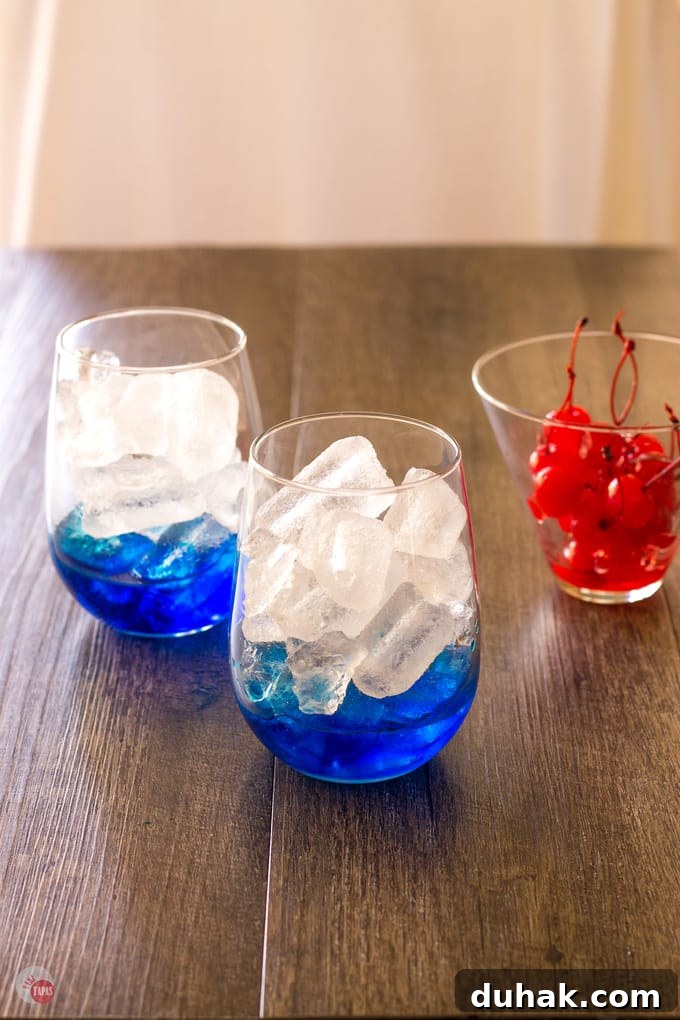 blue curacao in the bottom of 2 cocktail glasses and a small cup of maraschino cherries on a wood surface