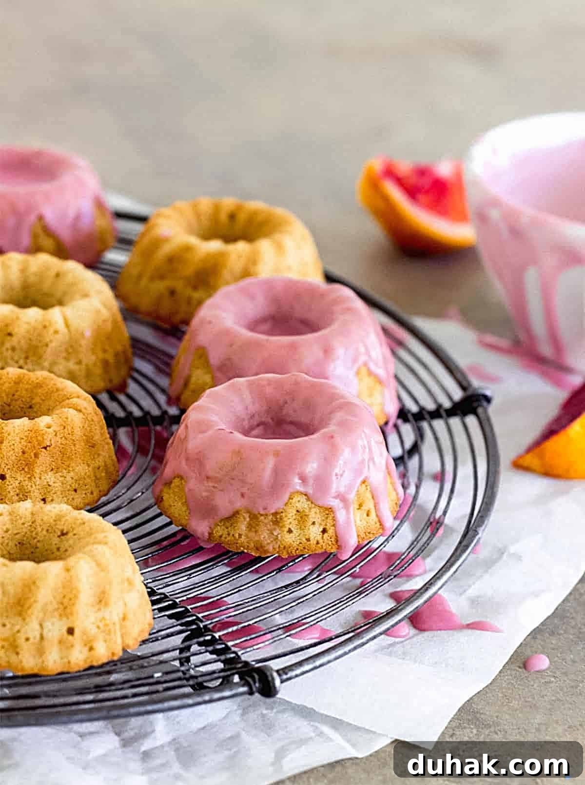 Pink Glazed mini bundt cakes on wire rack, grey surface, bowl and orange wedges on the side