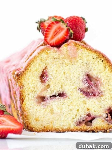 A cut loaf of strawberry pound cake, with a pink glaze and fresh strawberries. White background.