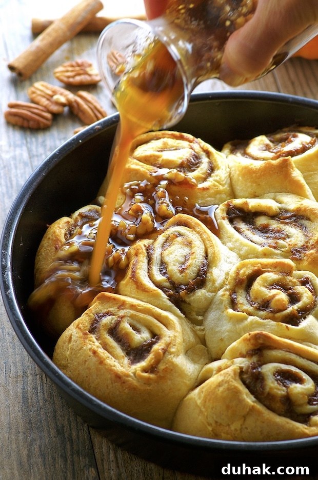 Maple-Banana Pumpkin Cinnamon Rolls with Warm Cinnamon Glaze 11 Side View of Easy Banana Pumpkin Cinnamon Rolls on a round black metal baking dish. A hand is holding a clear glass bottle with maple syrup and pecans pouring it over the pumpkin cinnamon rolls. On the background you there are some pecan a couple sticks of cinnamon