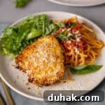 A plate with crispy air fryer parmesan crusted chicken, spaghetti, tomato sauce, and salad on a gray countertop.