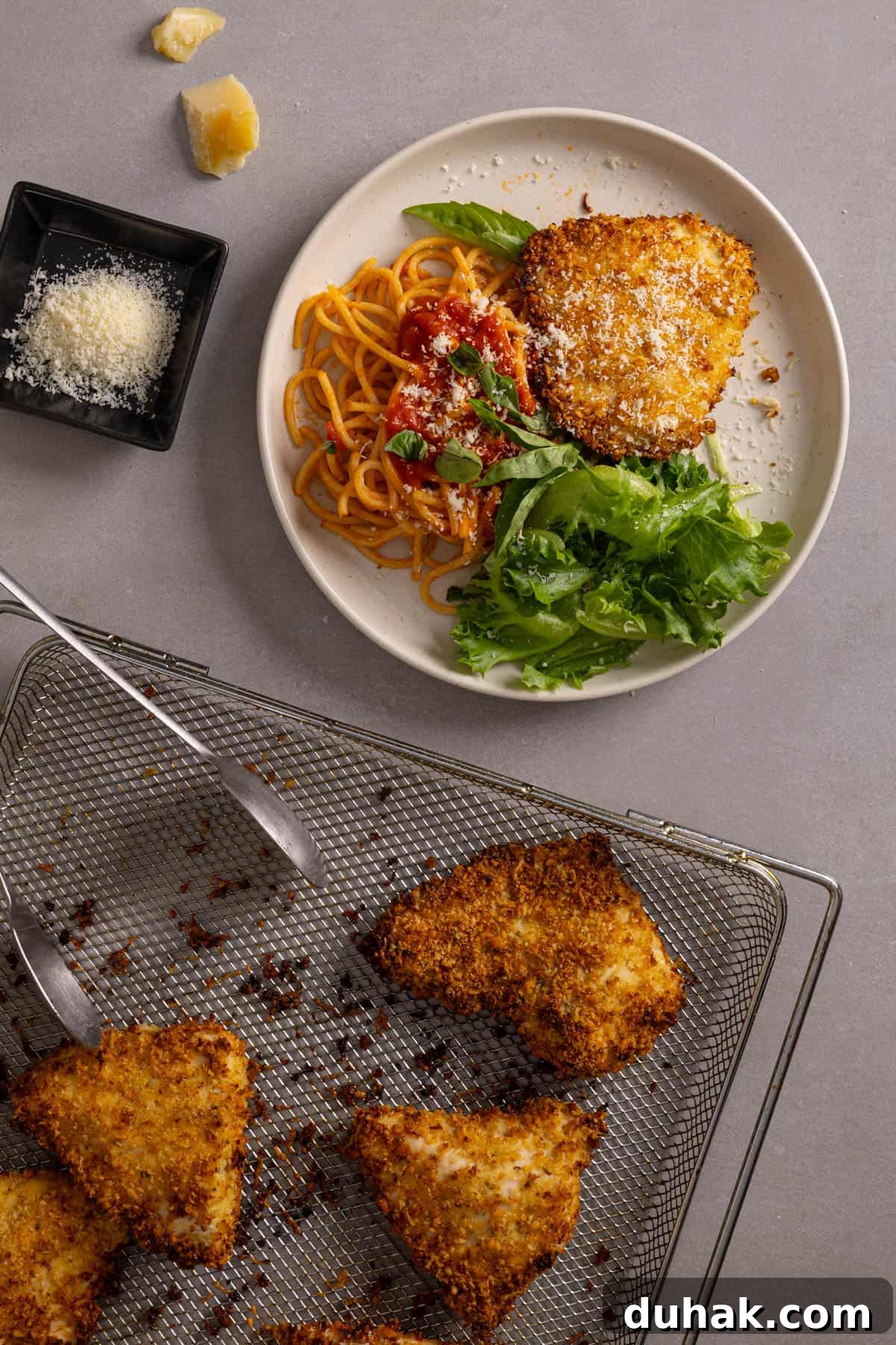 An air fryer basket with parmesan chicken and a plate with a serving of the chicken to the side.