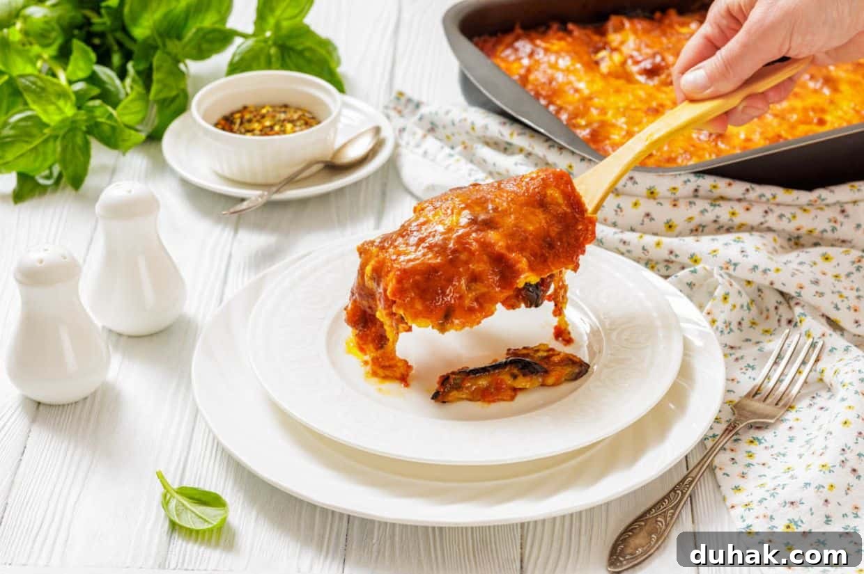 37 Comfort Food Recipes That Keep Reappearing on Menus 11 A hand serves a slice of Eggplant Parmigiana onto a white plate, with a casserole dish, herbs, and condiments visible on a white table.