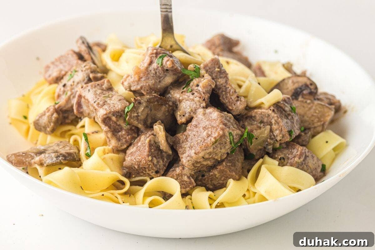 37 Comfort Food Recipes That Keep Reappearing on Menus 12 A white bowl filled with cooked egg noodles and chunks of beef, garnished with herbs, with a fork standing upright in the center.