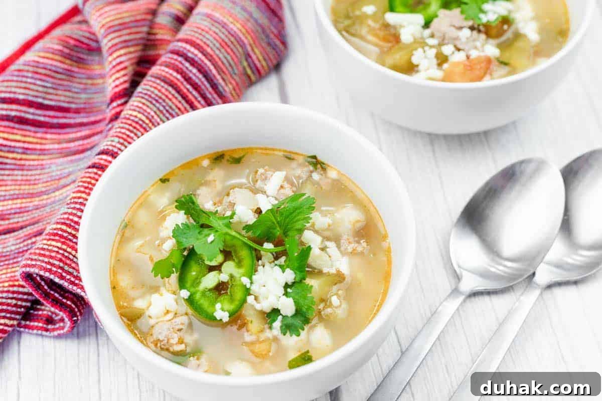 37 Comfort Food Recipes That Keep Reappearing on Menus 13 A bowl of Easy Green Pork Posole with white hominy, ground meat, sliced jalapeños, cheese, and cilantro, next to a striped cloth napkin and two metal spoons on a white surface.