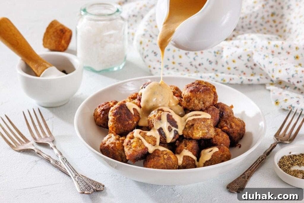 37 Comfort Food Recipes That Keep Reappearing on Menus 3 A bowl of meatballs is topped with creamy sauce being poured from a jug, surrounded by forks and seasoning jars on a white table.