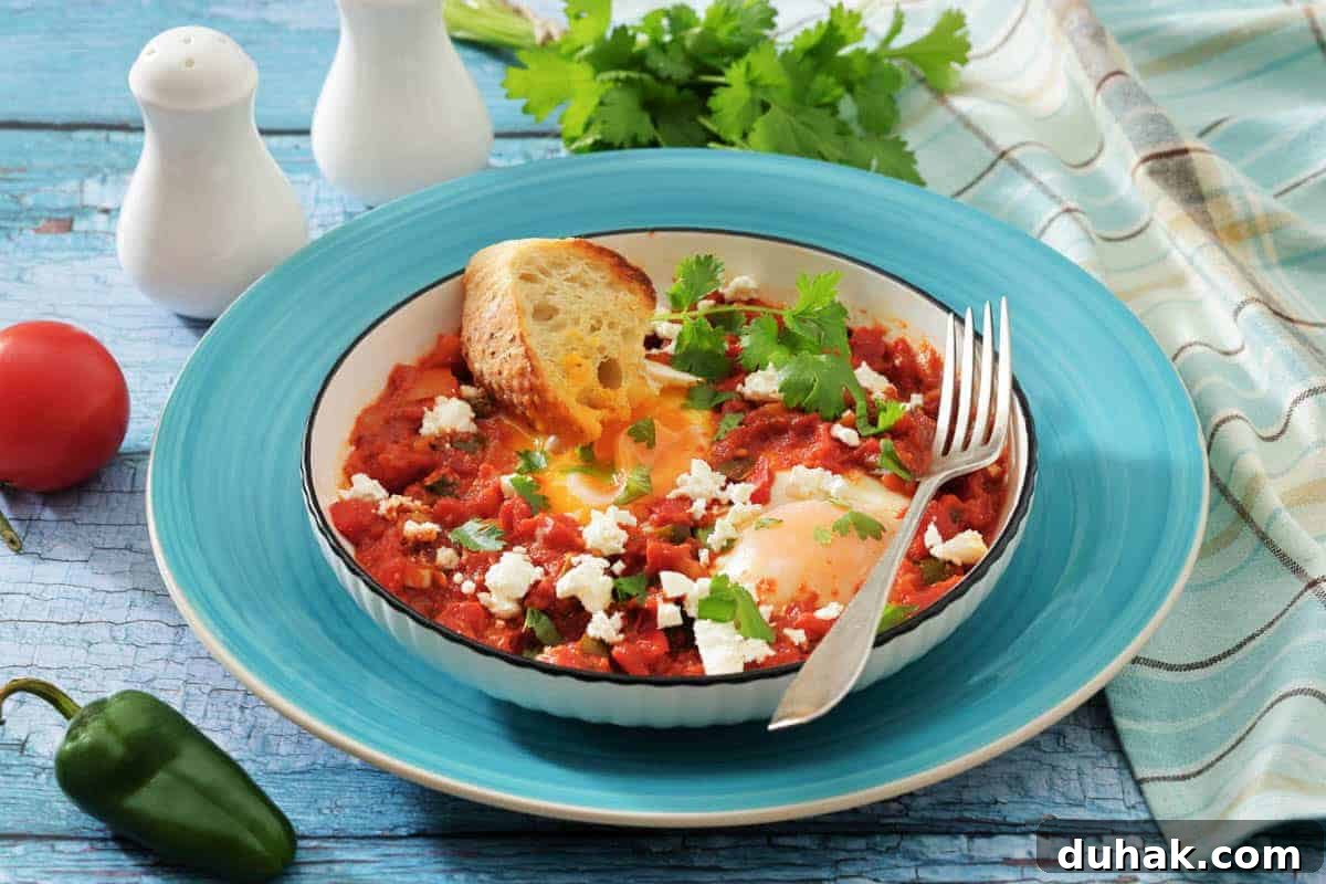 37 Comfort Food Recipes That Keep Reappearing on Menus 21 A bowl of shakshuka with poached eggs in tomato sauce, topped with feta and cilantro, served with a slice of toasted bread and a fork on a blue plate.