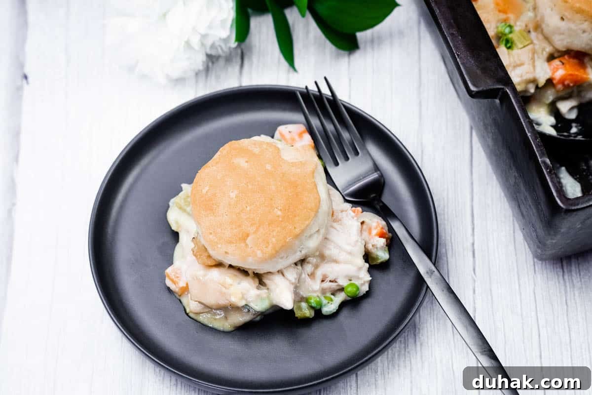 37 Comfort Food Recipes That Keep Reappearing on Menus 22 A black plate features a biscuit-topped serving of chicken pot pie, complete with chicken, peas, and carrots. A fork is placed on the plate, with a baking dish partially visible in the background.