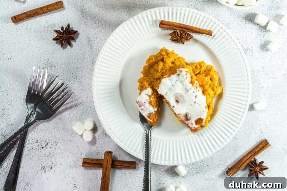 37 Comfort Food Recipes That Keep Reappearing on Menus 31 A plate with a serving of sweet potato casserole topped with marshmallows, surrounded by forks, marshmallows, cinnamon sticks, and star anise on a white surface.