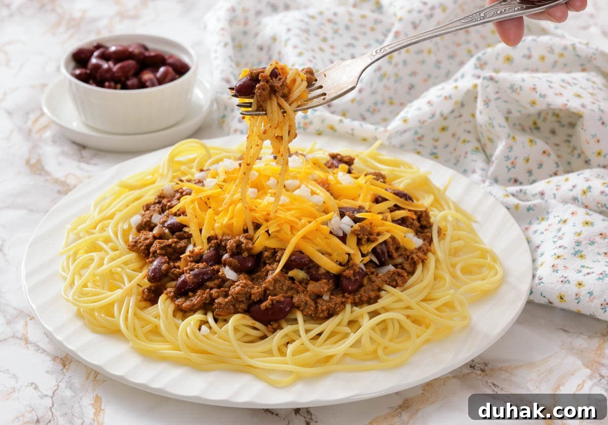 37 Comfort Food Recipes That Keep Reappearing on Menus 37 A plate of spaghetti topped with Cincinnati chili, shredded cheese, and beans, with a fork lifting a portion.