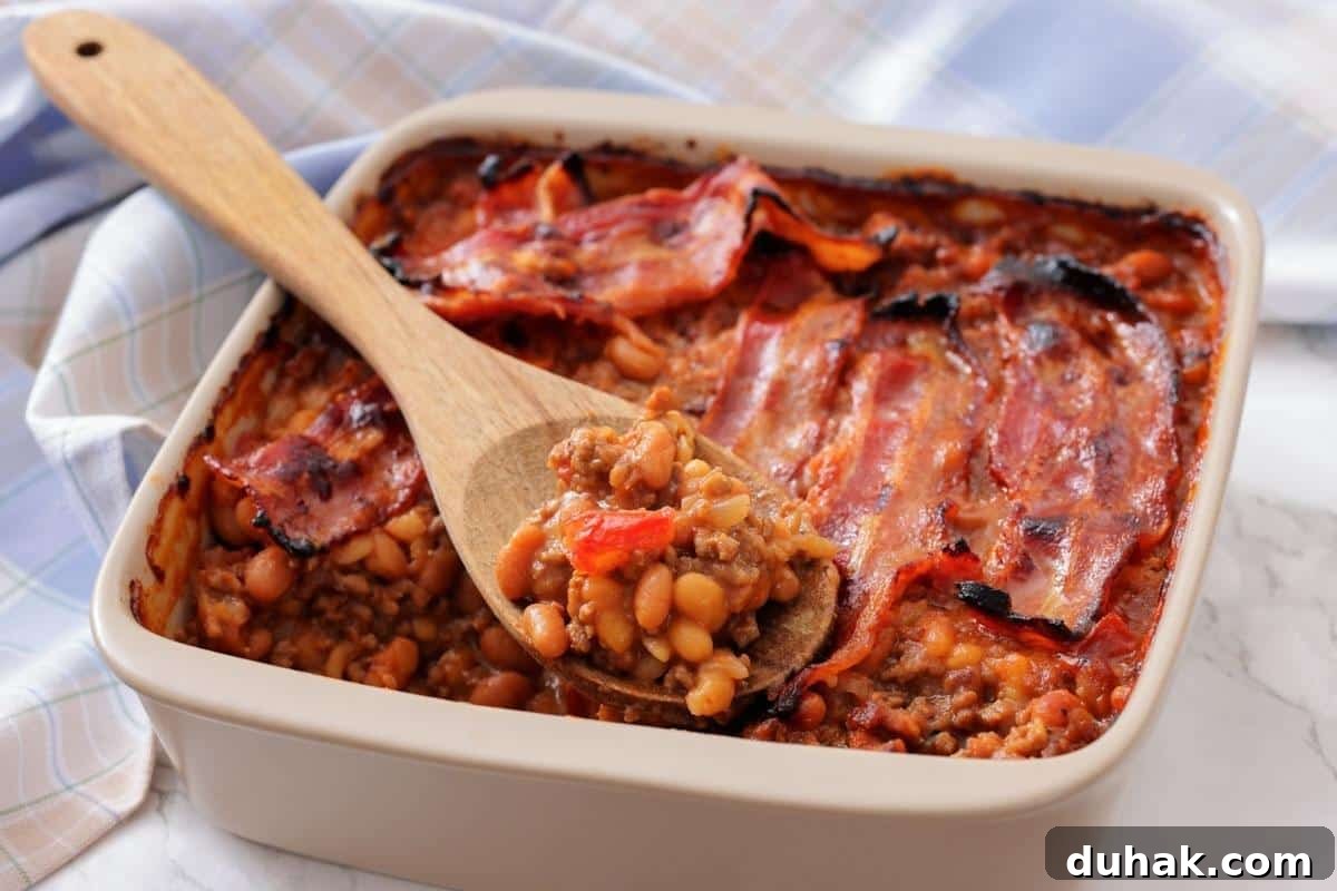 37 Comfort Food Recipes That Keep Reappearing on Menus 39 A square baking dish filled with Southern Ground Beef & Baked Bean Casserole and topped with strips of cooked bacon; a wooden spoon holds a serving.