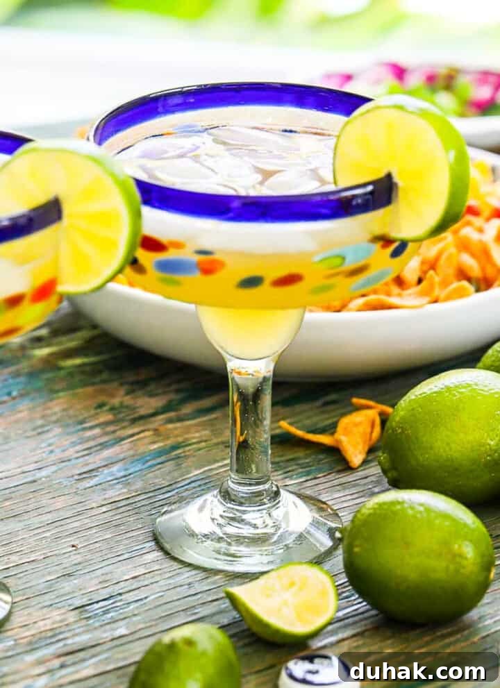 Blue rimmed margarita glasses with margaritas in them on ice cubes with sliced lime garnish.