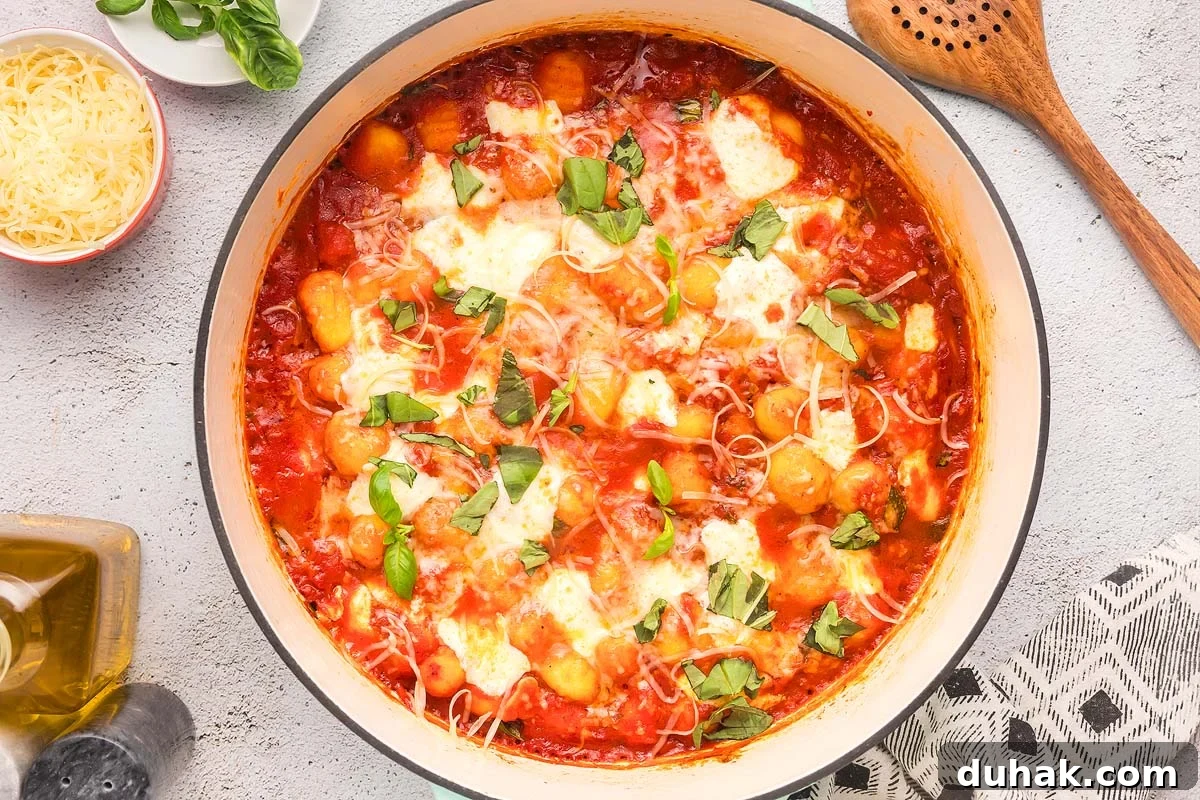 Sorrento-Style Gnocchi with Tomato, Basil & Mozzarella 2 A pot of baked gnocchi alla sorrentina in tomato sauce topped with melted cheese and fresh basil. Utensils and ingredients like grated cheese, olive oil, and more fresh basil are nearby.