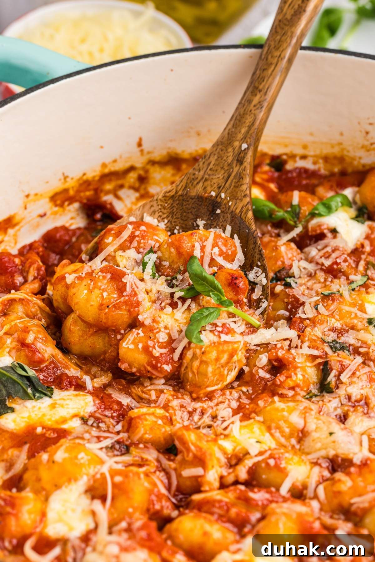 Sorrento-Style Gnocchi with Tomato, Basil & Mozzarella 11 Close-up of a wooden spoon stirring gnocchi alla sorrentina in tomato sauce, topped with grated cheese and garnished with basil, inside a white pot.