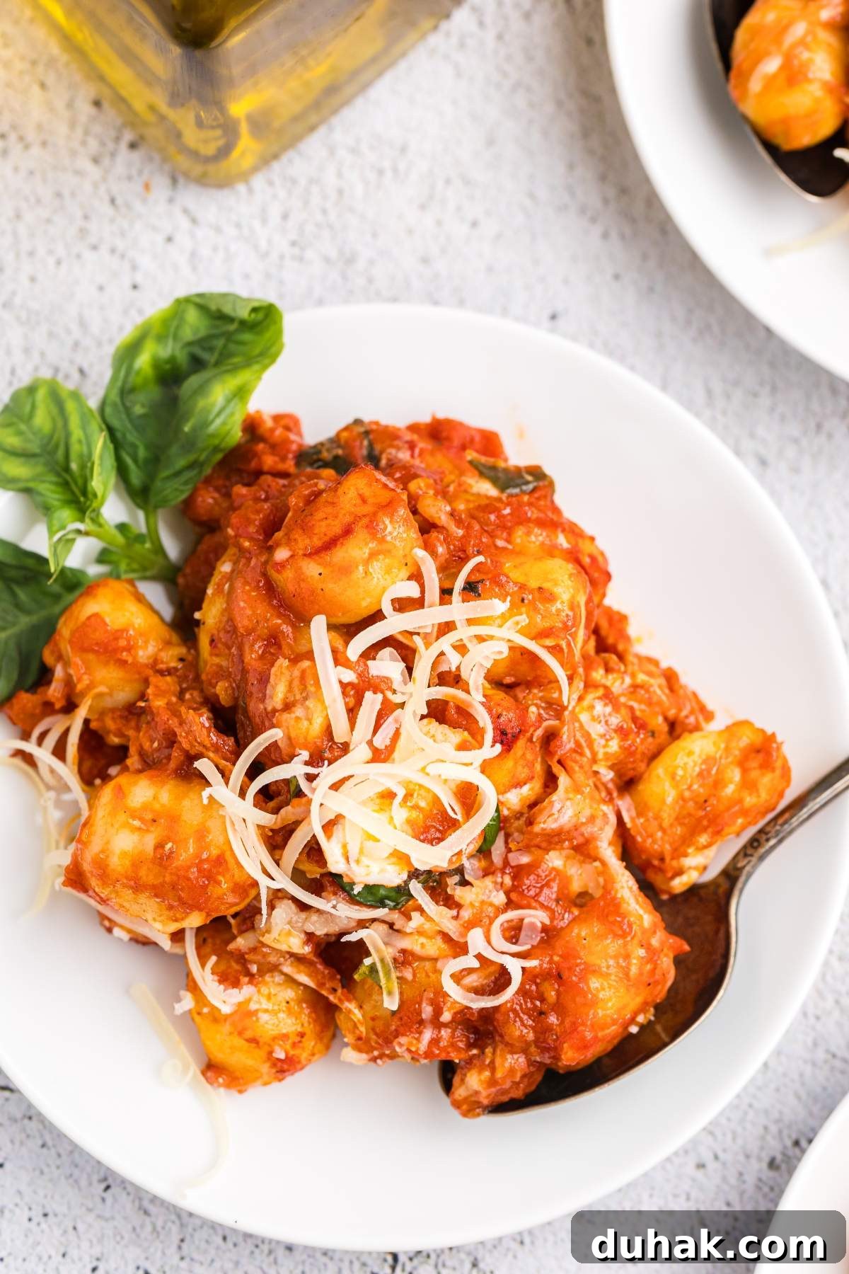 Sorrento-Style Gnocchi with Tomato, Basil & Mozzarella 12 A white plate contains gnocchi alla sorrentina, bathed in rich tomato sauce and garnished with shredded cheese and basil. A spoon rests on the side of the plate.