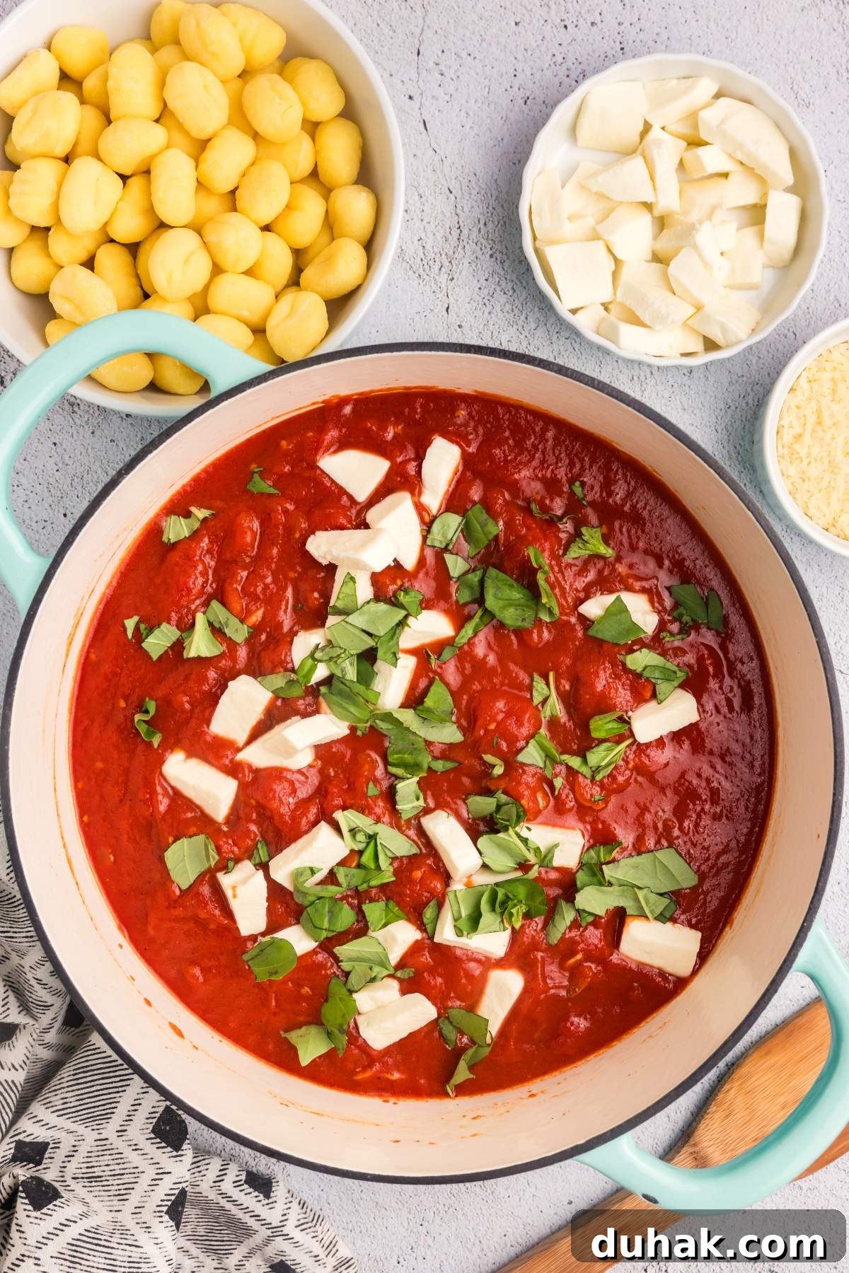 Sorrento-Style Gnocchi with Tomato, Basil & Mozzarella 16 Top-down view of a saucepan containing tomato sauce topped with chopped fresh basil and cheese chunks, embodying the essence of gnocchi alla sorrentina. Surrounding the pan are bowls of uncooked gnocchi, cheese pieces, and grated cheese.