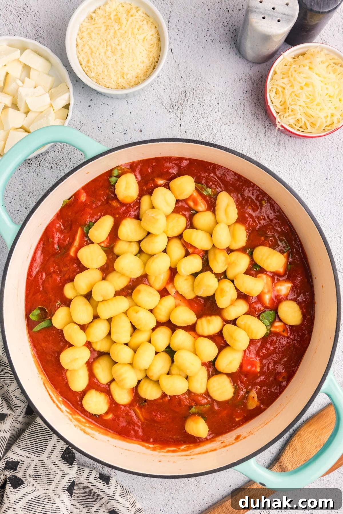Sorrento-Style Gnocchi with Tomato, Basil & Mozzarella 17 A pot of gnocchi alla sorrentina in red tomato sauce is accompanied by bowls of grated cheese and diced mozzarella. A wooden spoon rests nearby on a patterned cloth.