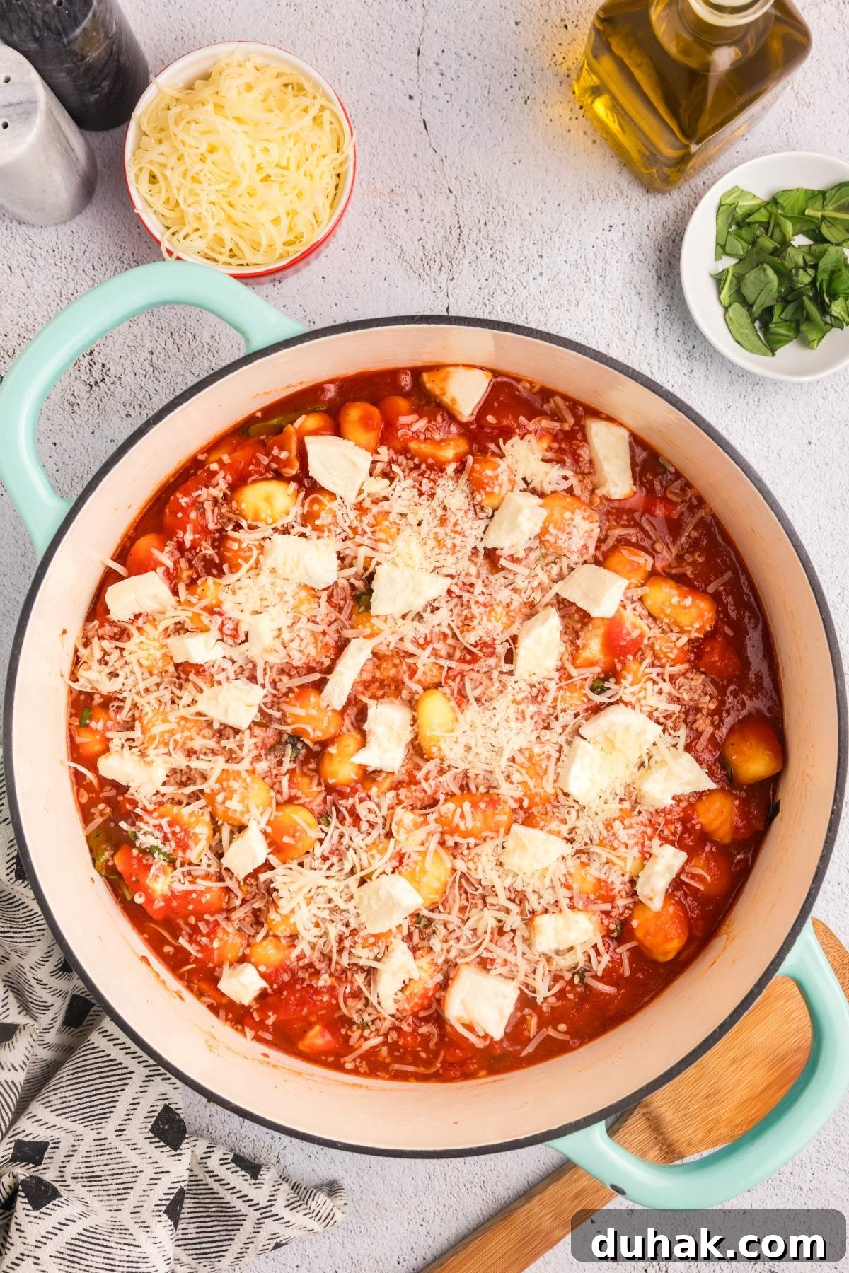 Sorrento-Style Gnocchi with Tomato, Basil & Mozzarella 18 A large pot of gnocchi alla sorrentina (sorrentina style gnocchi) in a tomato sauce, topped with melted cheese. Surrounding the pot are bowls of shredded cheese and fresh basil, along with an olive oil bottle and a wooden spatula.