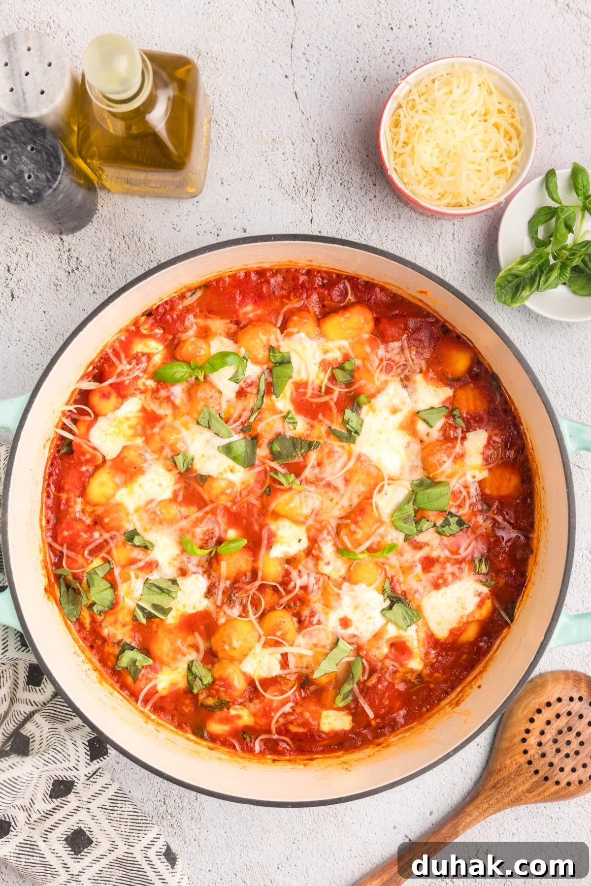 Sorrento-Style Gnocchi with Tomato, Basil & Mozzarella 19 A pot of gnocchi alla sorrentina (gnocchi sorrento style), topped with melted cheese and fresh basil, sits on a countertop. Nearby are olive oil, shredded cheese in a bowl, pepper, and fresh basil leaves.
