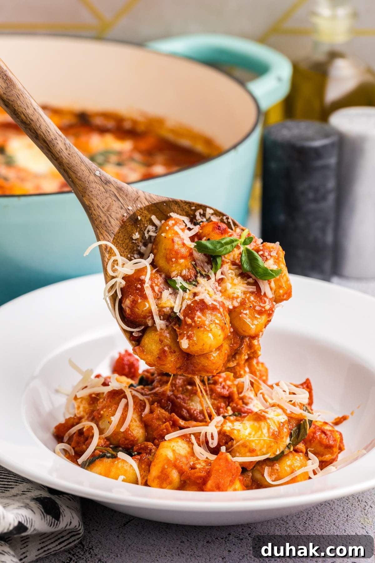 Sorrento-Style Gnocchi with Tomato, Basil & Mozzarella 5 A wooden spoon holding gnocchi alla sorrentina in tomato sauce over a white bowl, topped with grated cheese and basil, with a pot in the background.