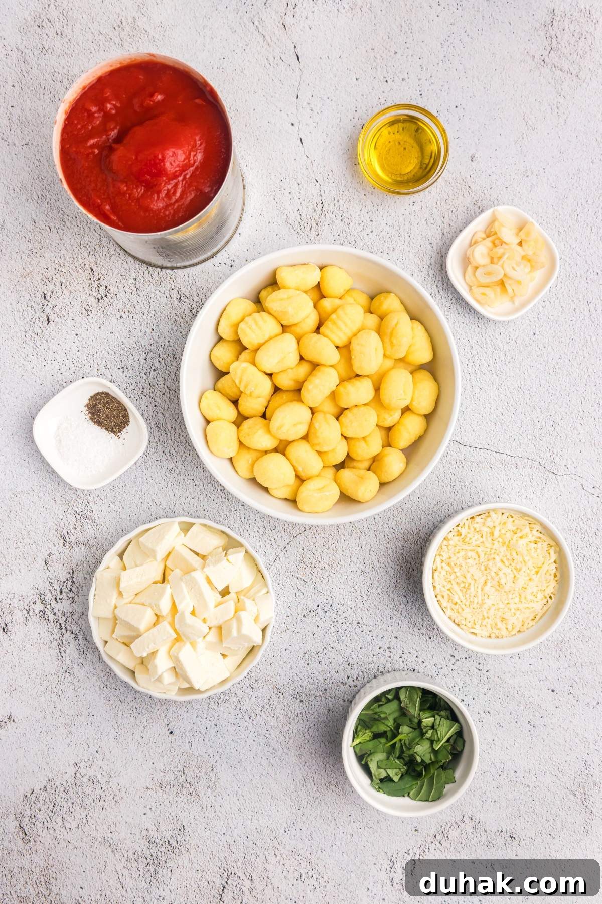 Sorrento-Style Gnocchi with Tomato, Basil & Mozzarella 6 Top-down view of a table with bowls of ingredients to make gnocchi alla sorrentina. Chopped mozzarella, parmesan, sliced garlic, fresh basil, salt and pepper, a can of tomato sauce, and a small bowl of olive oil.