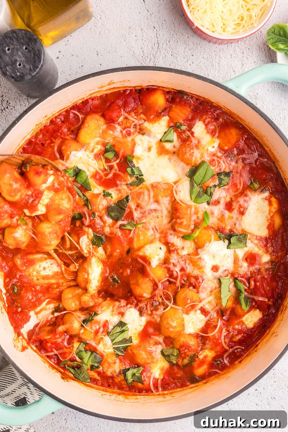 Sorrento-Style Gnocchi with Tomato, Basil & Mozzarella 10 A pot of gnocchi sorrento in tomato sauce topped with melted cheese and fresh basil leaves, next to a pepper shaker, a bottle of oil, and a small bowl of grated cheese on a light-colored surface.