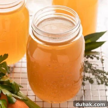 Golden chicken bone broth in glass jars with carrots, celery, and parsley, cooling on the counter.