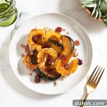 A plate of roasted winter squash topped with cranberry agrodolce sauce and hazelnuts, a green glass are in the upper left, a bowl of fresh sage is in the upper right, a gold fork is in the lower right.