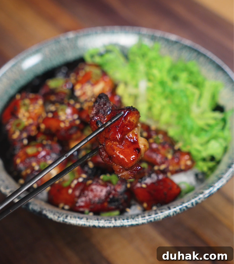 A juicy piece of garlic soy glazed chicken is held up with chopsticks over a bowl filled with caramelized chicken thighs, fluffy rice, and fresh shredded lettuce. The glossy, sticky glaze clings to the chicken, highlighting its crispy edges and tender inside, making the bite look irresistible and packed with flavor.