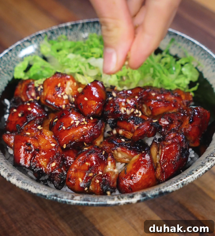 A hand sprinkles sesame seeds over a freshly cooked bowl of garlic soy glazed chicken. The chicken pieces are deeply caramelized with crisp edges and glistening glaze, sitting atop white rice with a side of crisp lettuce, creating the perfect final touch for texture and visual appeal.