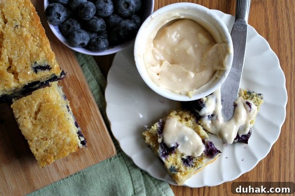 overhead shot of blueberry cornbread on a cutting board with a bowl of fresh blueberries and a slice of cornbread on a white plate with butter on it and a bowl of butter and a knife