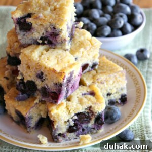 pieces of blueberry cornbread stacked on a plate with a bowl of blueberries in the background- set on a green towel on a wooden surface