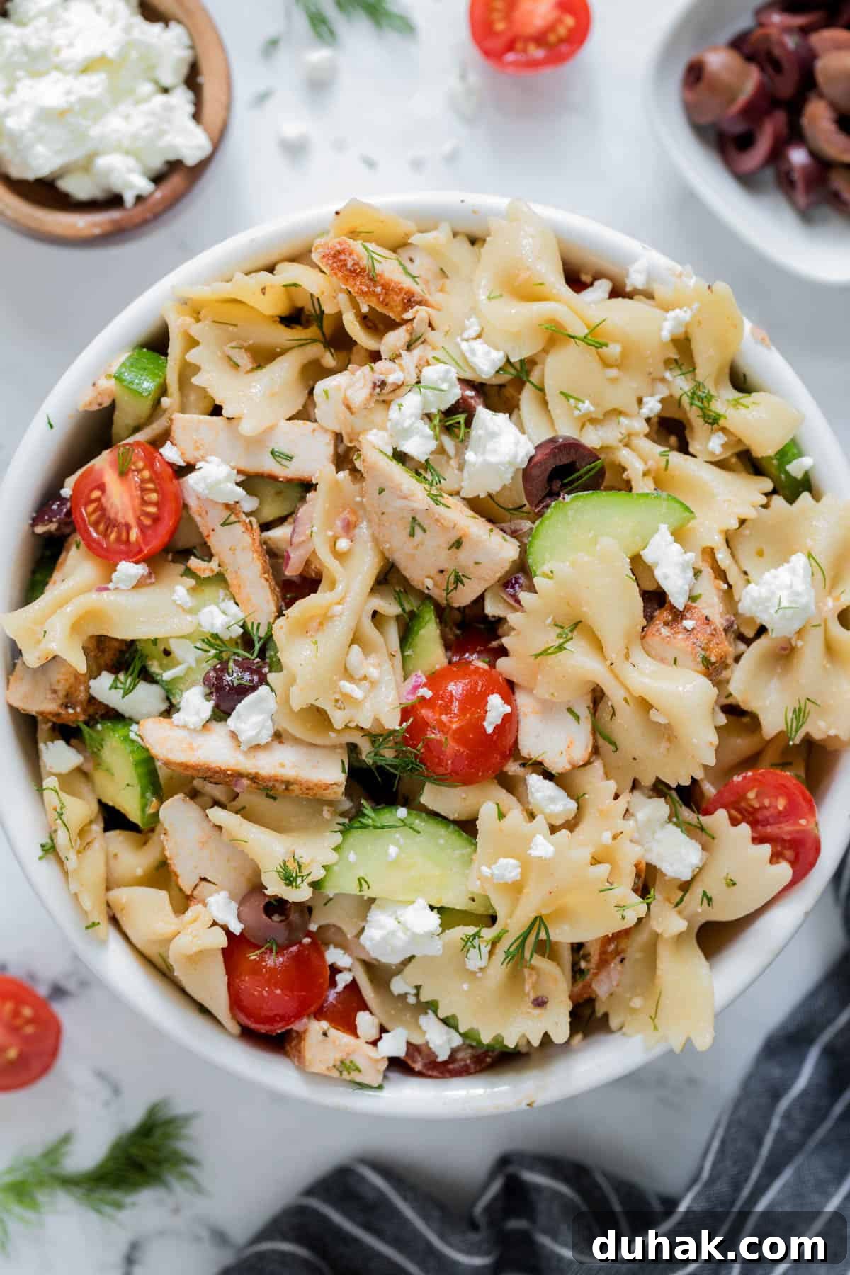Greek Chicken Pasta Salad with Feta & Lemon Vinaigrette 2 Greek chicken pasta salad in a white bowl for serving. Cucumbers, tomatoes, kalamata olives, cooked chicken, bowtie pasta and feta cheese are piled high in the bowl.