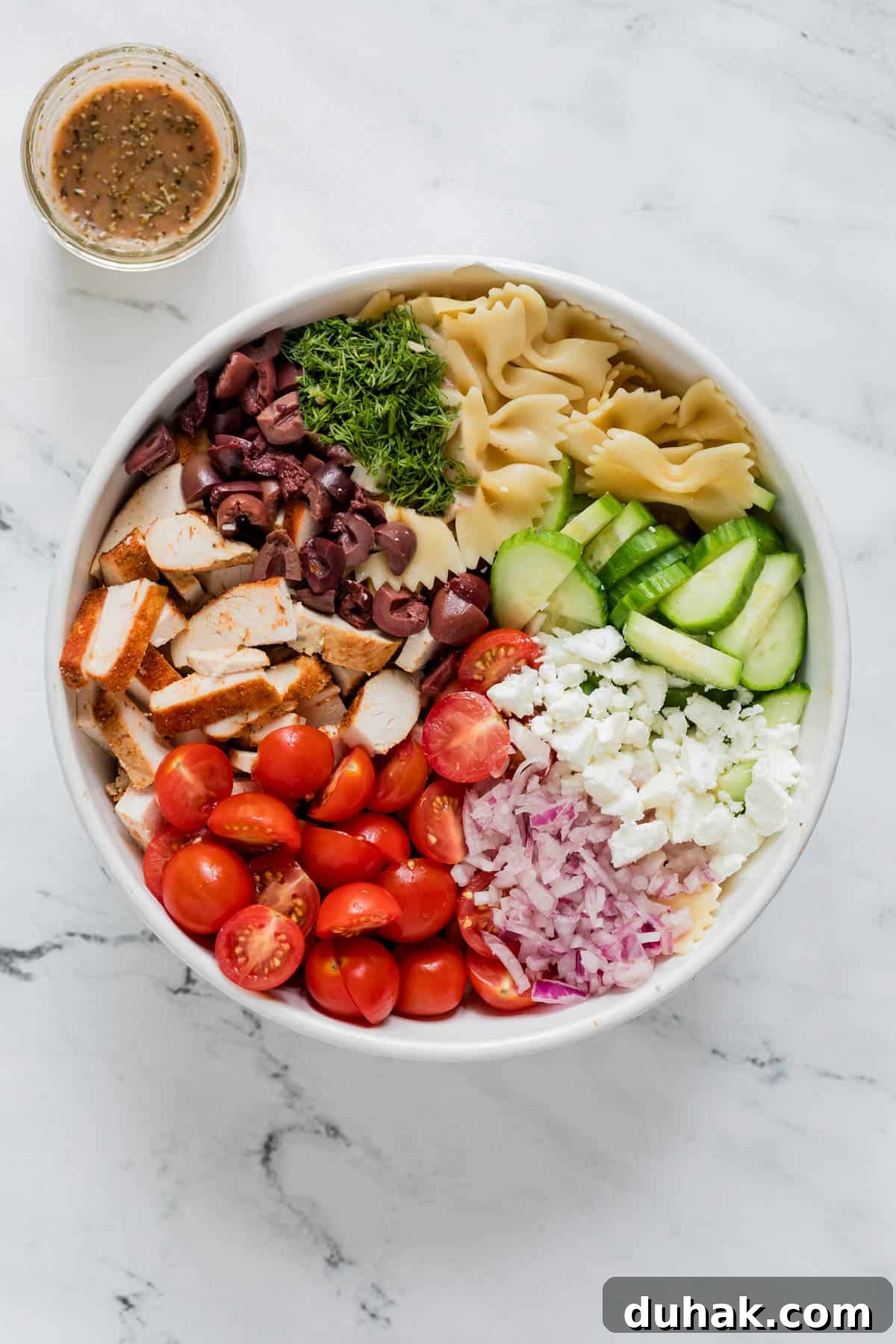 Greek Chicken Pasta Salad with Feta & Lemon Vinaigrette 3 Ingredients for the greek chicken salad sit in a white bowl before mixing including cooked bow tie pasta, cherry tomatoes, cucumber, kalamata olives, red onion, feta cheese and fresh dill. A small glass with the homemade vinaigrette sits on the side.