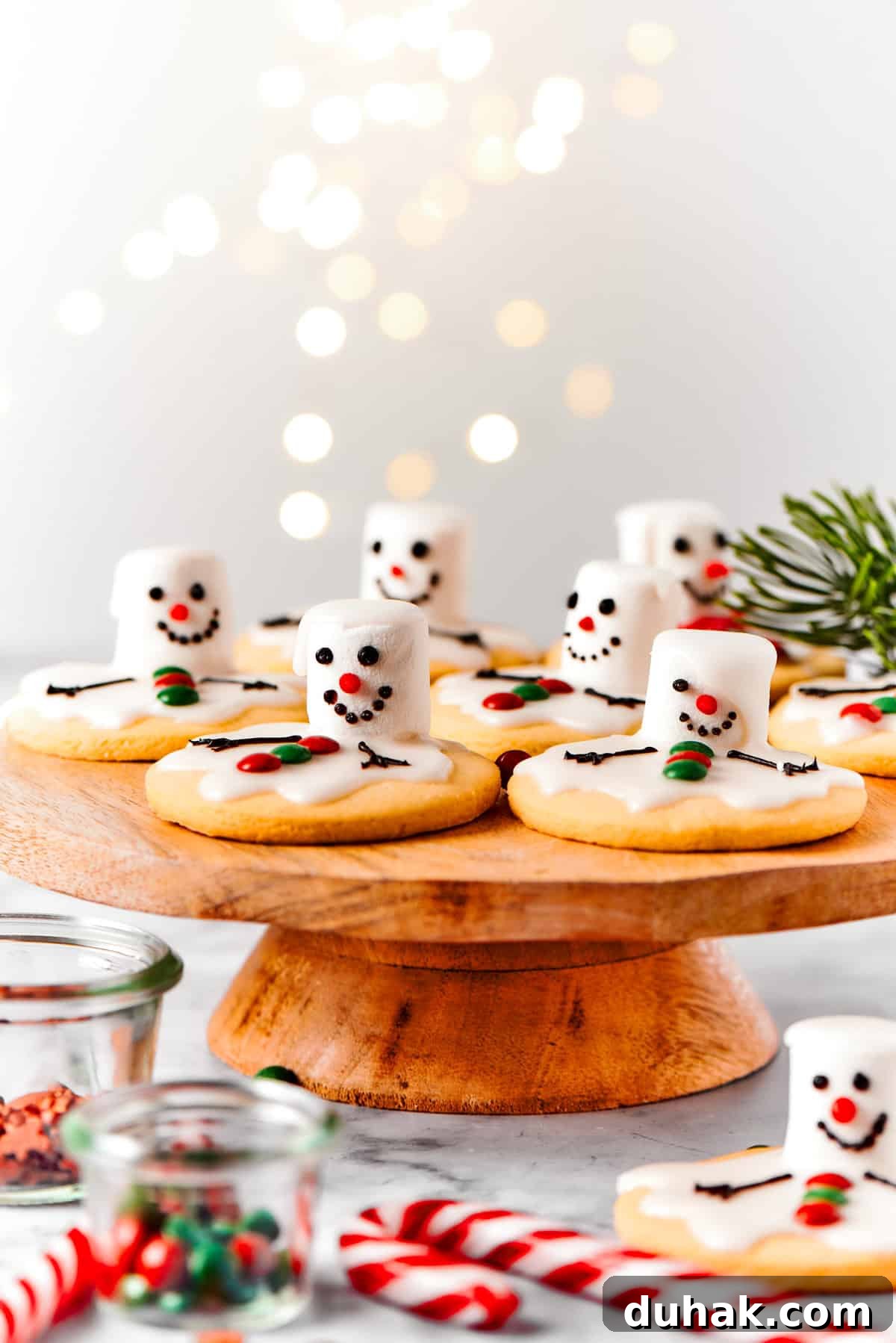 Melting snowman cookies are arranged on a wooden cake plate, with holiday decorations nearby.