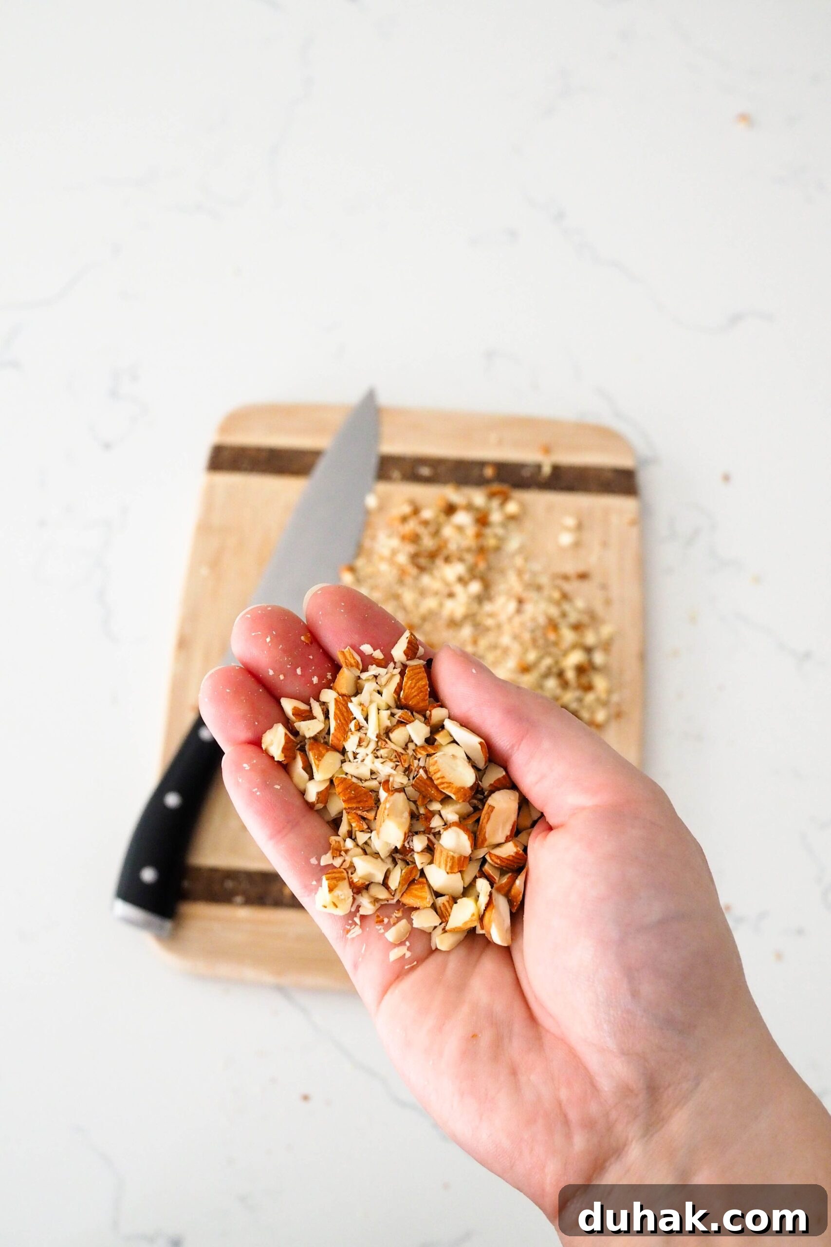 A hand holds up chopped almonds in front of a cutting board and knife.