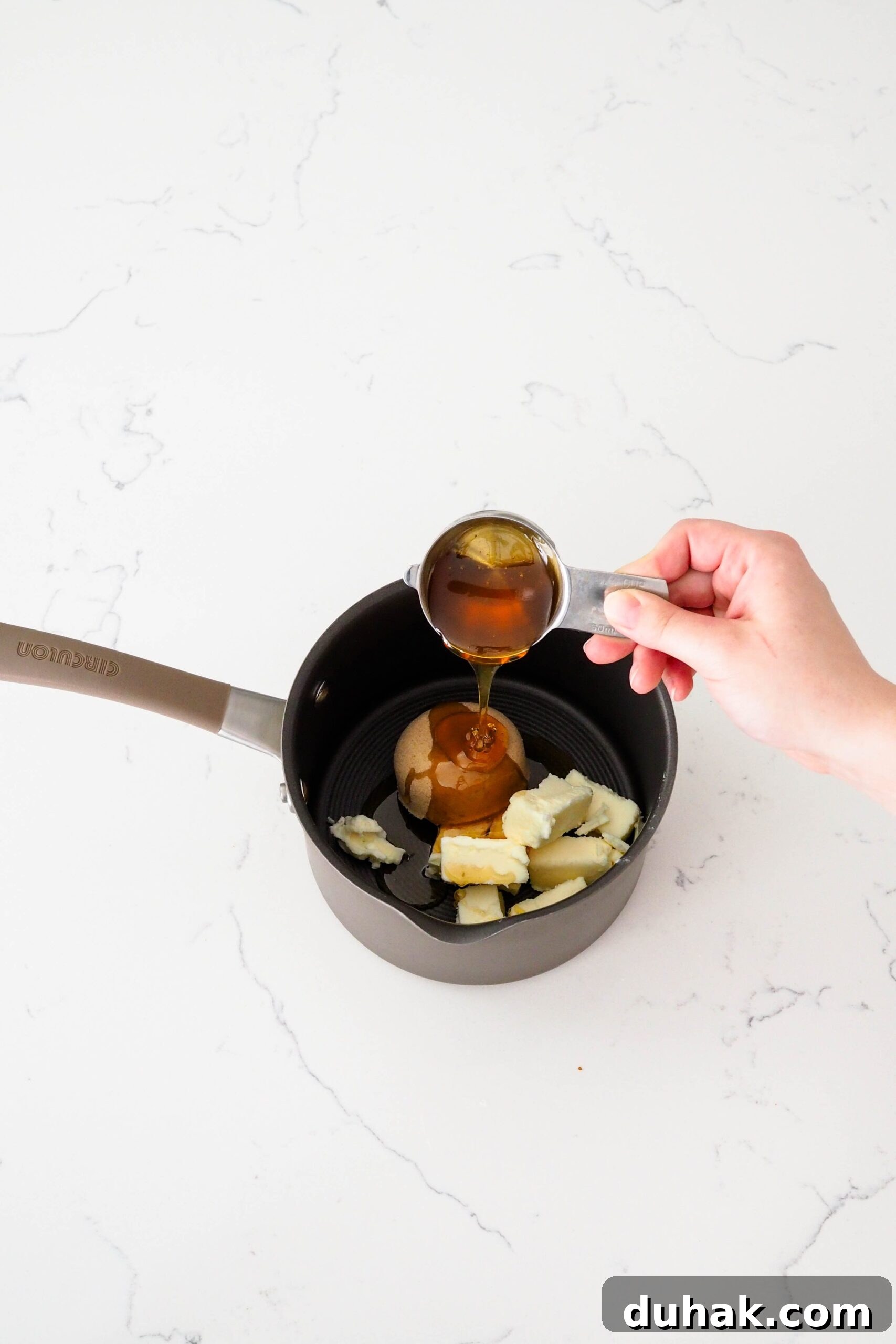 A hand pours honey into a medium pot with butter and brown sugar.