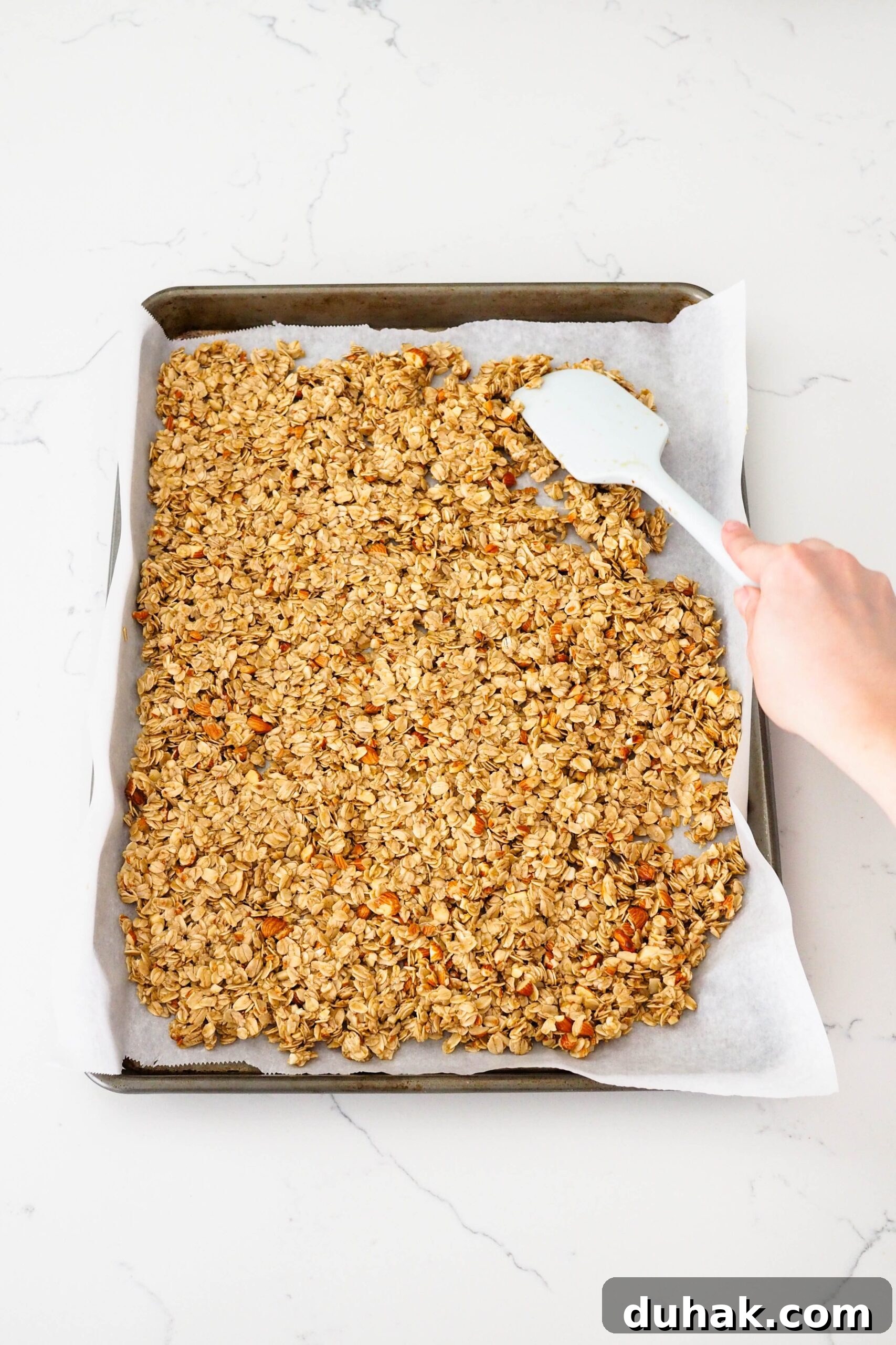 A spatula spreads out granola on a baking sheet.