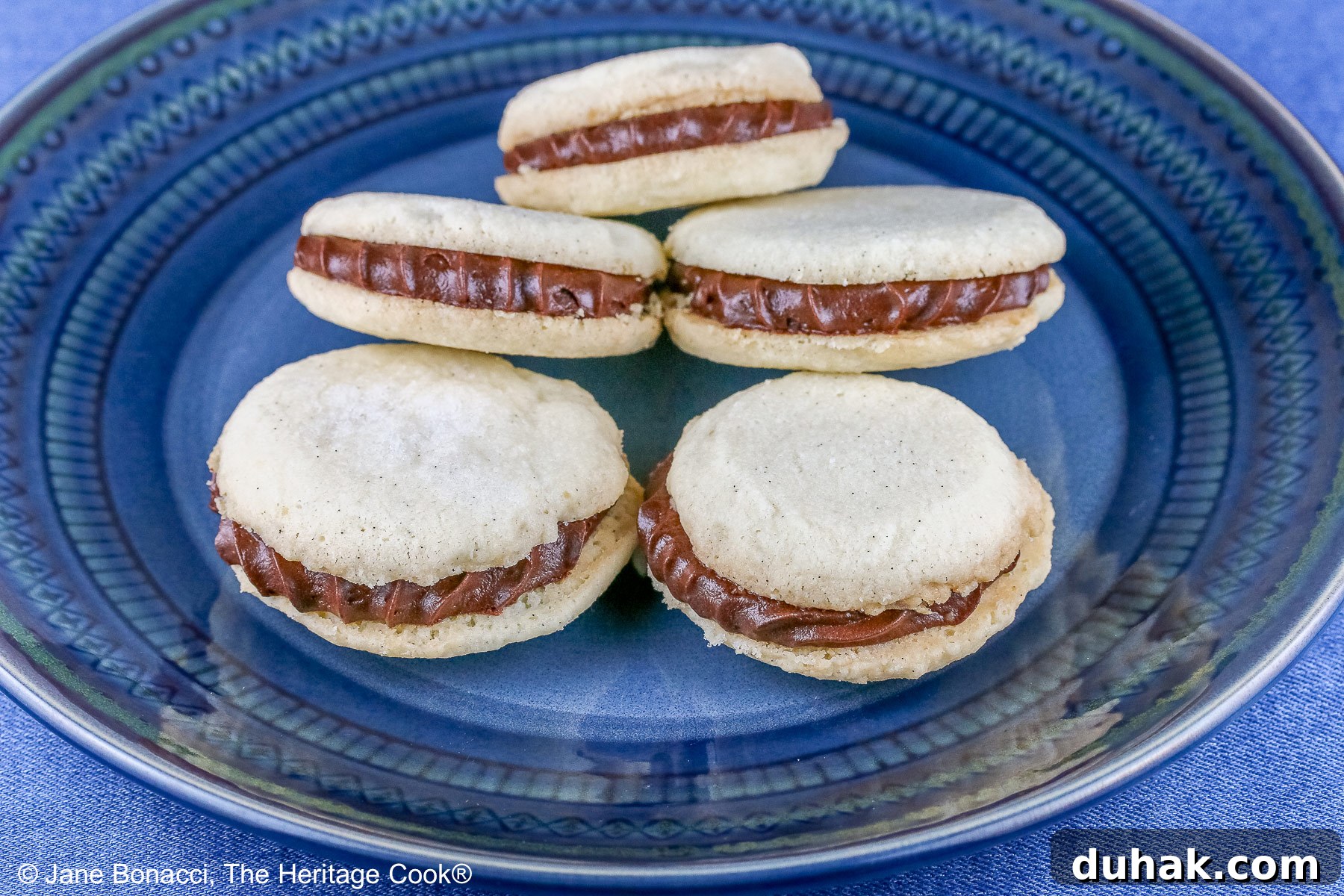 Stack of Chocolate Stuffed Vanilla Shortbread Cookies on either a white plate or blue plate on a bold blue background, the cookies staggered in a pile, the filling piped on with a star tip © 2025 Jane Bonacci, The Heritage Cook.