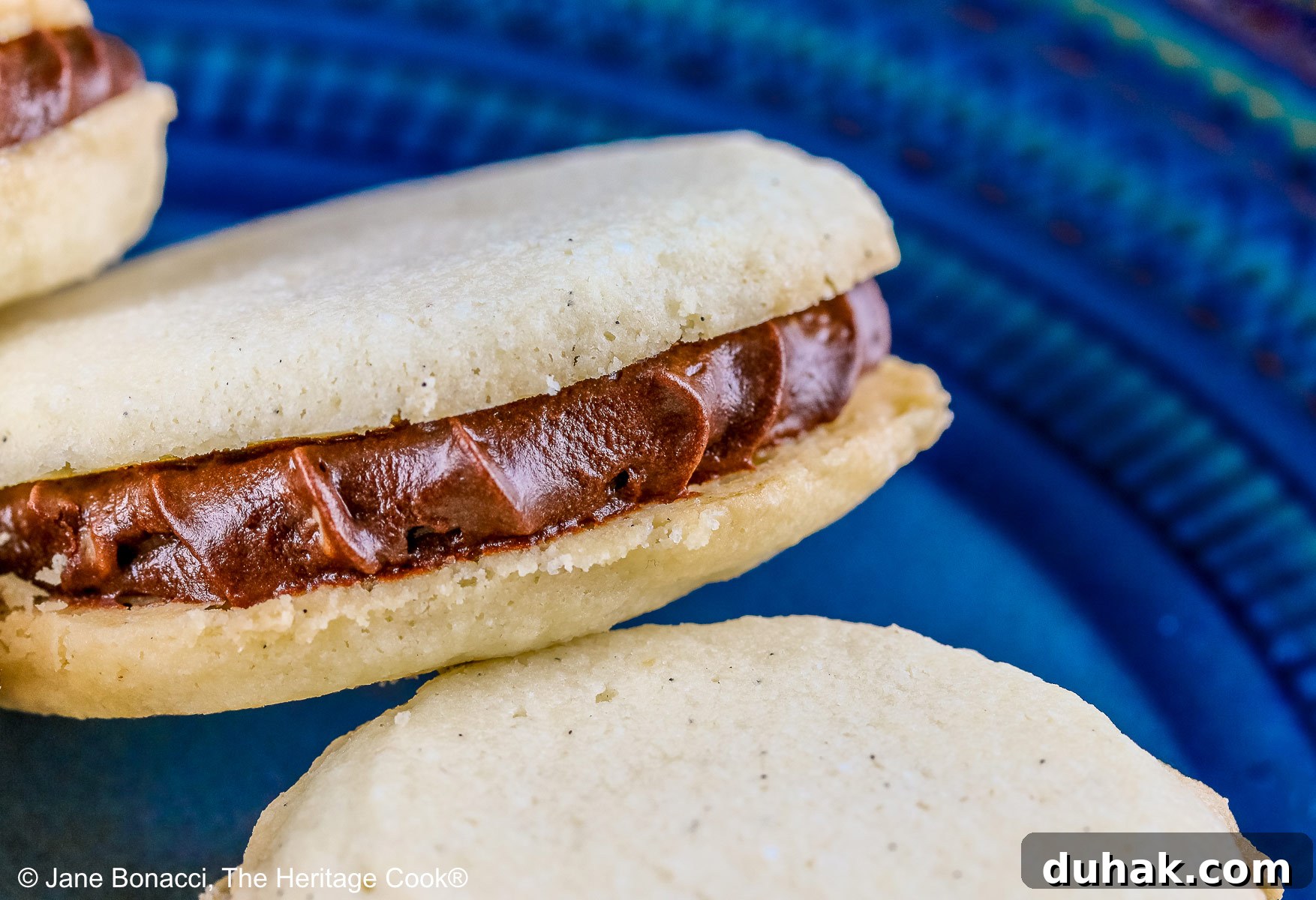 Stack of Chocolate Stuffed Vanilla Shortbread Cookies on either a white plate or blue plate on a bold blue background, the cookies staggered in a pile, the filling piped on with a star tip © 2025 Jane Bonacci, The Heritage Cook.