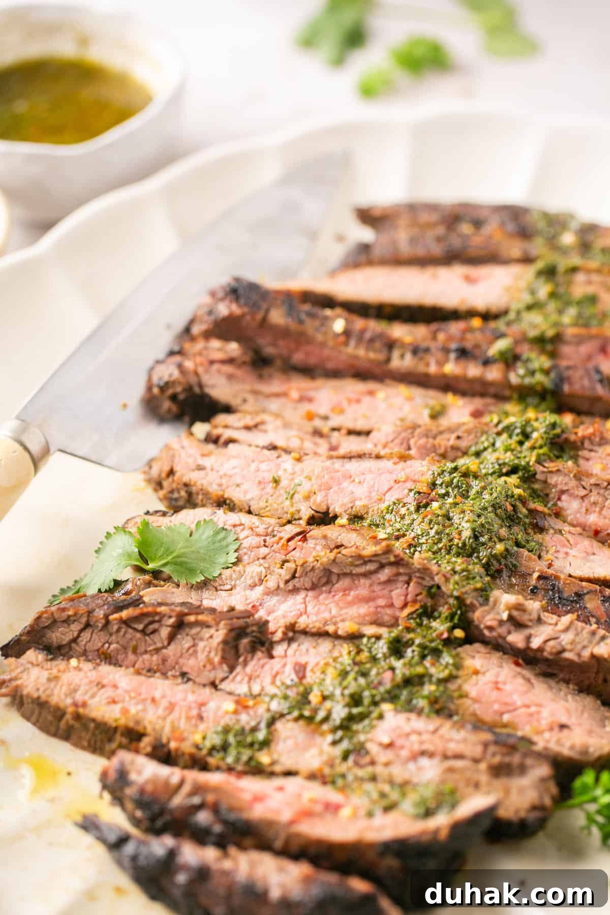 Cut flank steak on a white platter with chimichurri sauce on top of it with a bowl of chimichurri beside it.