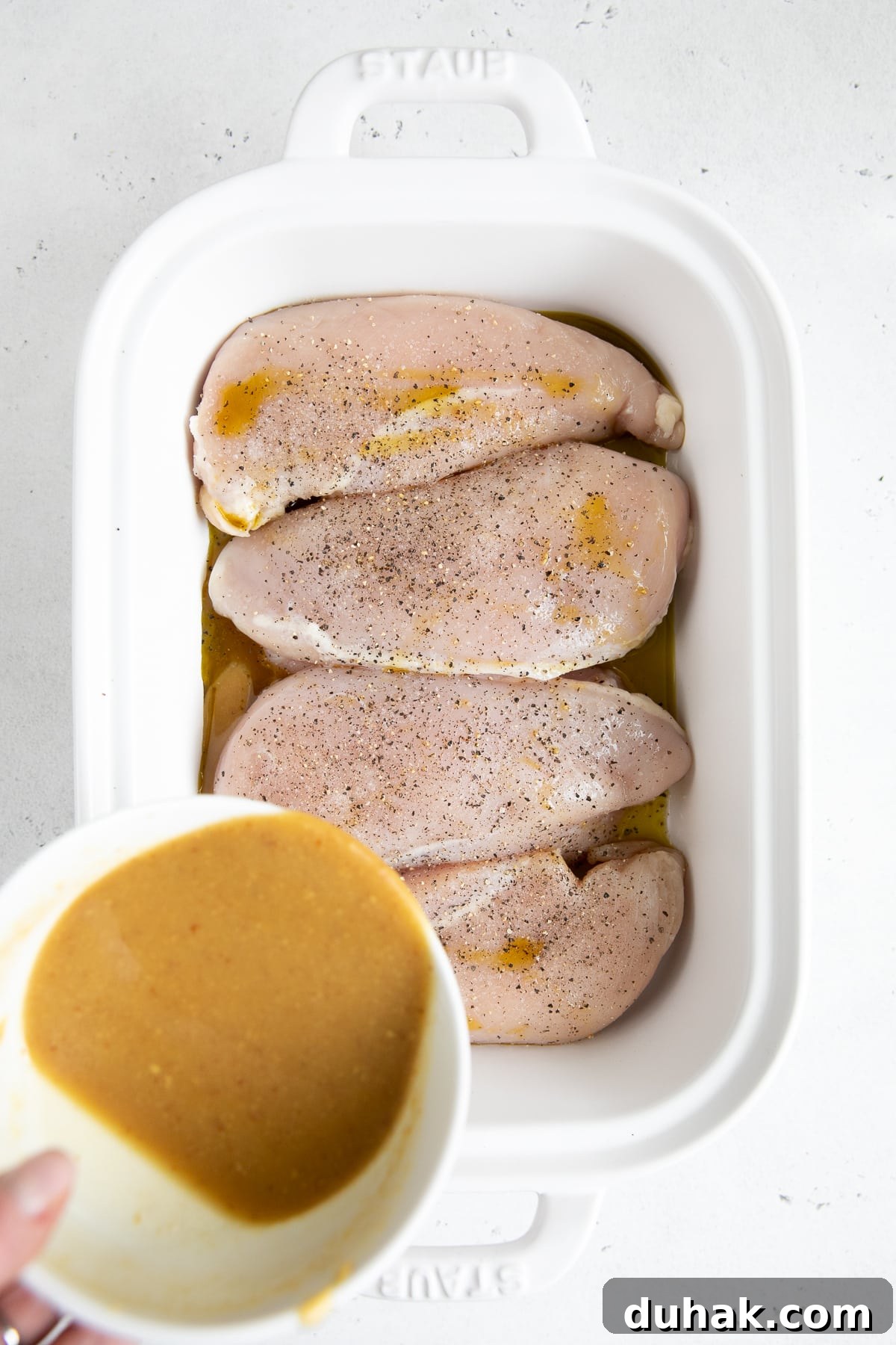 Four raw chicken breasts seasoned with salt and pepper in a white baking dish.
