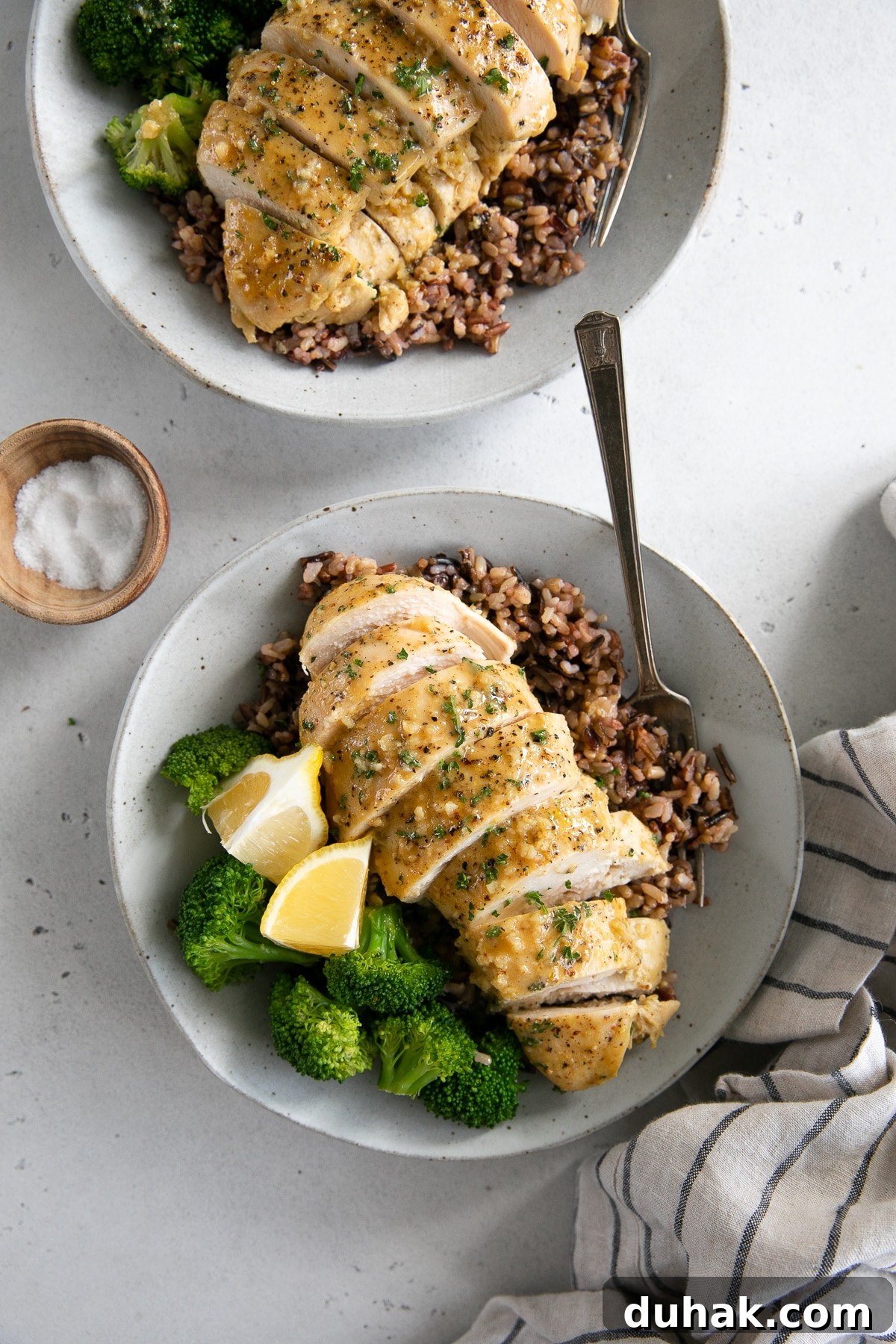 Two white plates with sliced honey mustard chicken breast over a bed of cooked rice and served with steamed broccoli and fresh lemon.