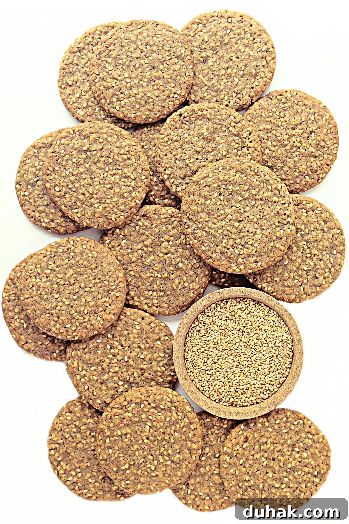 Brown sesame wafer cookies with a bowl of toasted sesame seeds on a white background.