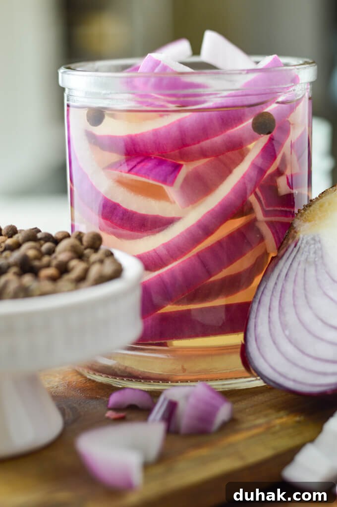 A jar of pickled onions sit on a wooden tray. Allspice berries and a half cut red onion are in the forefront of the photo.