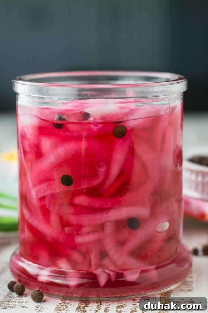 A jar of pickled red onions sit in a pink vinegar solution. Allspice berries float in the vinegar.