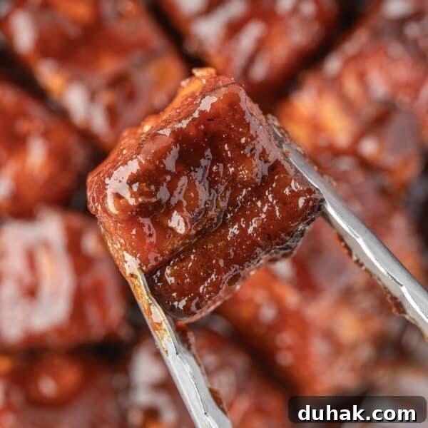 Close-up of metal tongs holding a glazed, smoked pork belly cube with more tender pork belly bites visible in the blurred background.