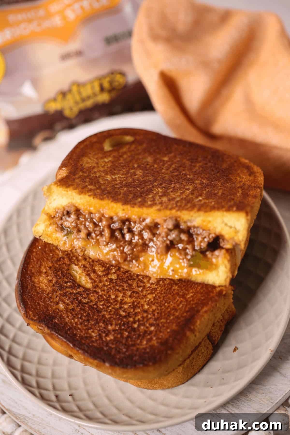 Sloppy Joe Grilled Cheese Sandwich Recipe for Comfort Dinner 6 A Sloppy Joe grilled cheese sandwich, with a filling of seasoned ground beef and gooey melted cheese, is partially sliced and artfully stacked on a plate. In the background, a bread package adds the finishing touch to this deliciously messy creation.