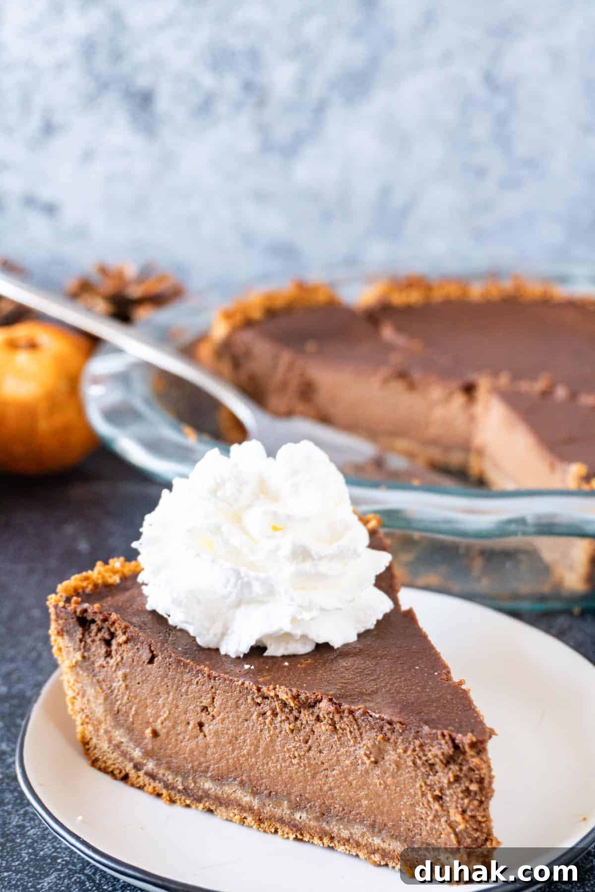Decadent Chocolate Pumpkin Pie Recipe for Fall Desserts 2 A slice of Chocolate Pumpkin Pie on a white plate, topped with whipped cream, with the rest of the pie in the background.