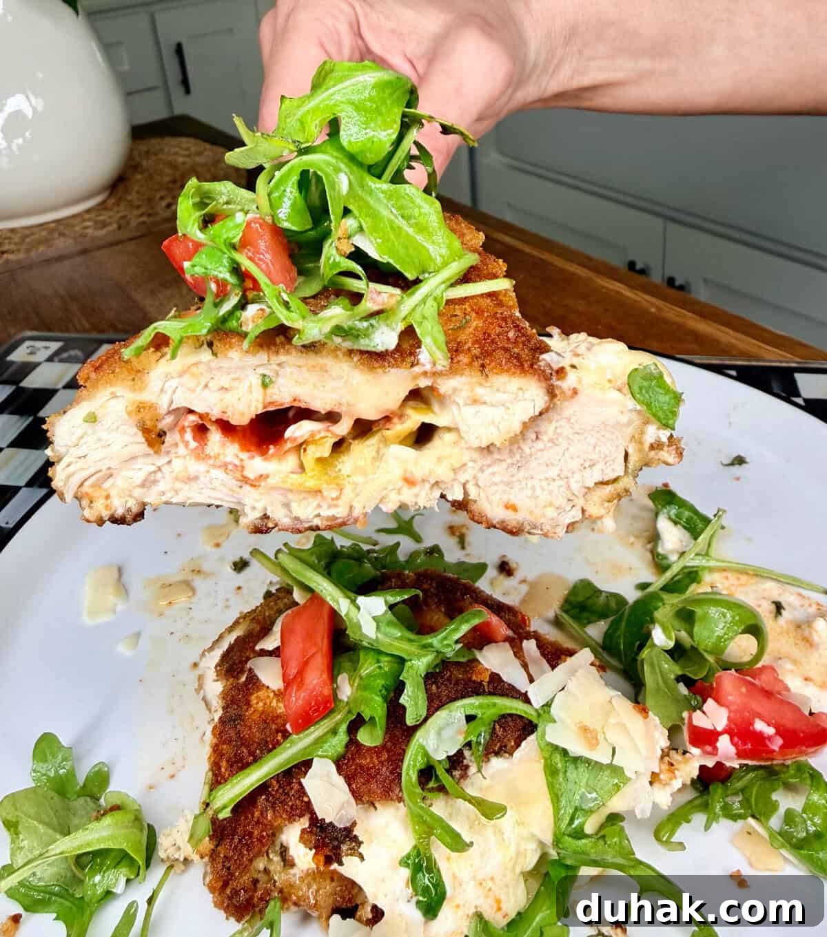 Creamy Spinach & Cheese Stuffed Chicken Cutlets Recipe 12 A hand holds a breaded, stuffed chicken breast topped with arugula, tomato, and shaved cheese above a plate with more of the same dish on it, in a bright kitchen setting.